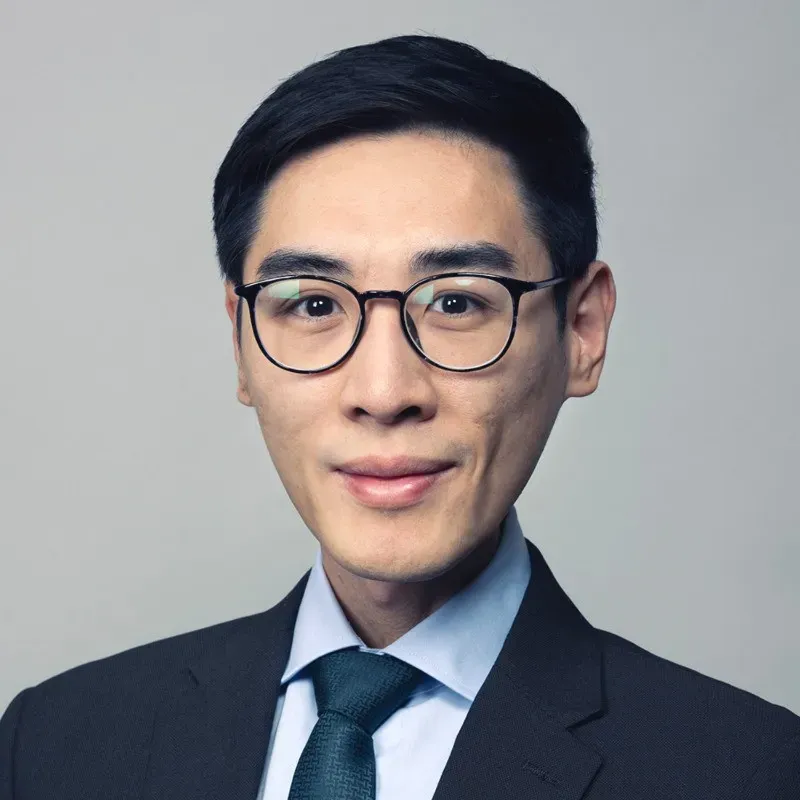 Portrait of a young man with short black hair, wearing round glasses, a dark suit, light blue shirt, and dark tie against a grey background.