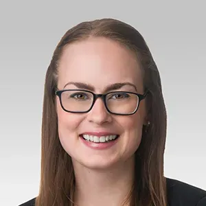Smiling woman with long brown hair and black-rimmed glasses against a light gray background.