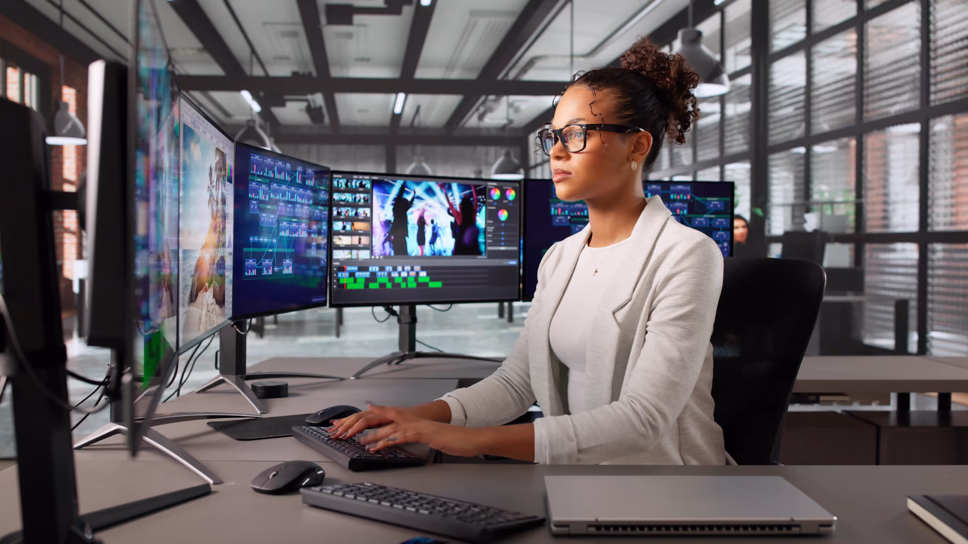 Woman with glasses editing video on a computer with multiple monitors in an office.