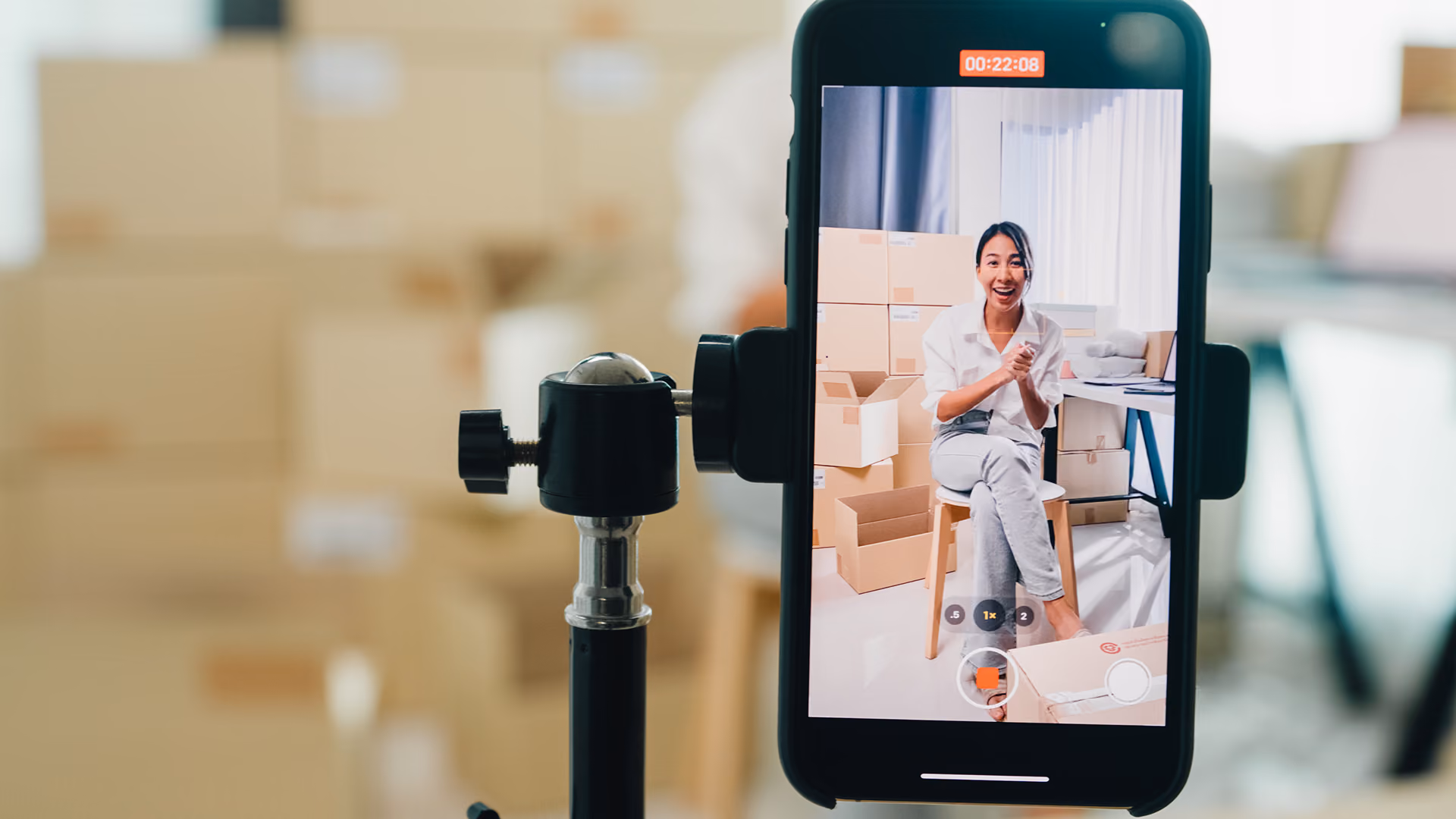 Woman recording video on phone sitting on stool surrounded by cardboard boxes in a bright room.