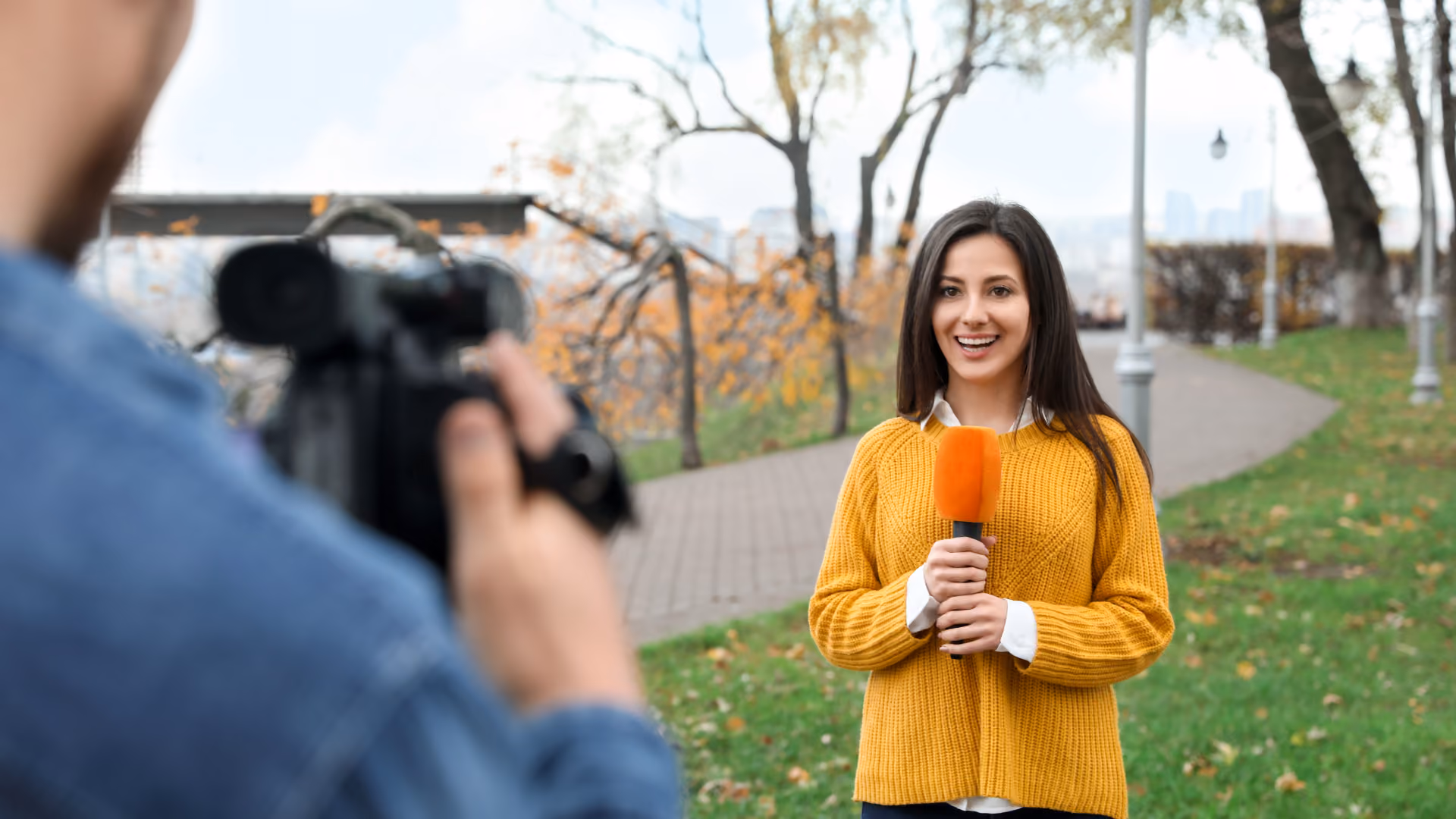 Smiling female reporter in yellow sweater holds orange microphone outdoors during video recording.