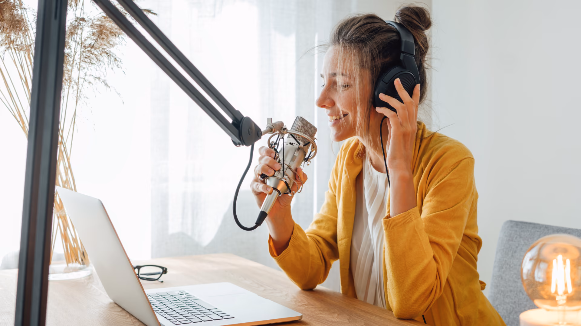 Woman in yellow cardigan recording with microphone wearing headphones in front of a laptop.