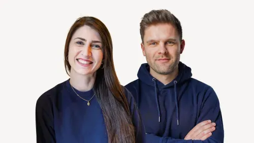 Fröhliche Frau mit langen braunen Haaren und goldener Halskette neben einem Mann mit kurzen braunen Haaren und blauem Hoodie mit verschränkten Armen.