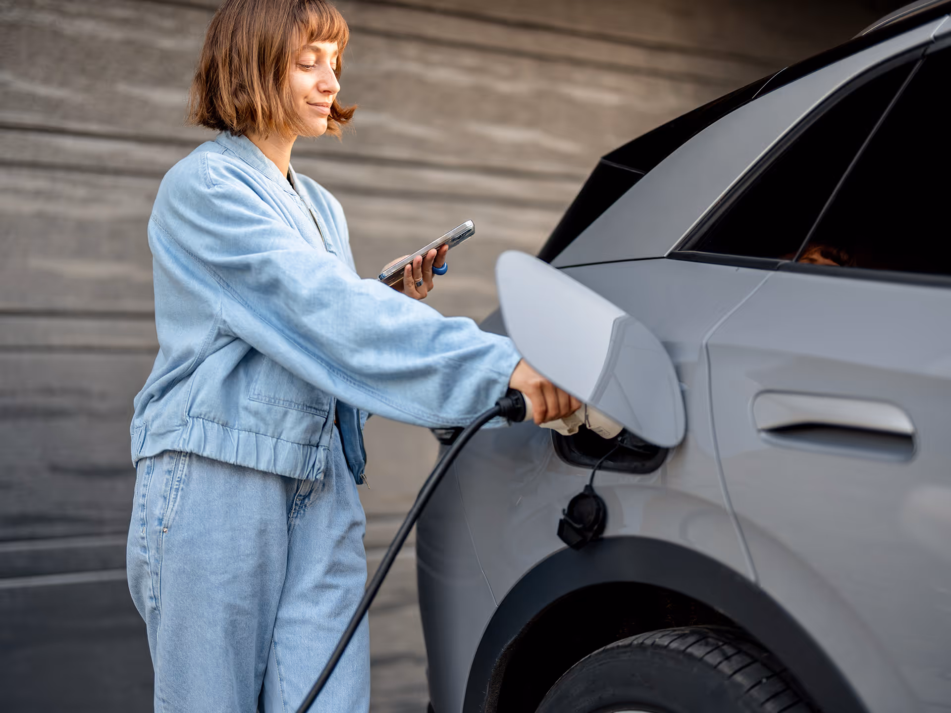 Frau lädt ein Elektroauto mit Ladekabel und hält dabei ein Smartphone in der Hand.