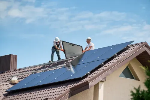 Zwei Arbeiter installieren Solarmodule auf einem Dach unter blauem Himmel.