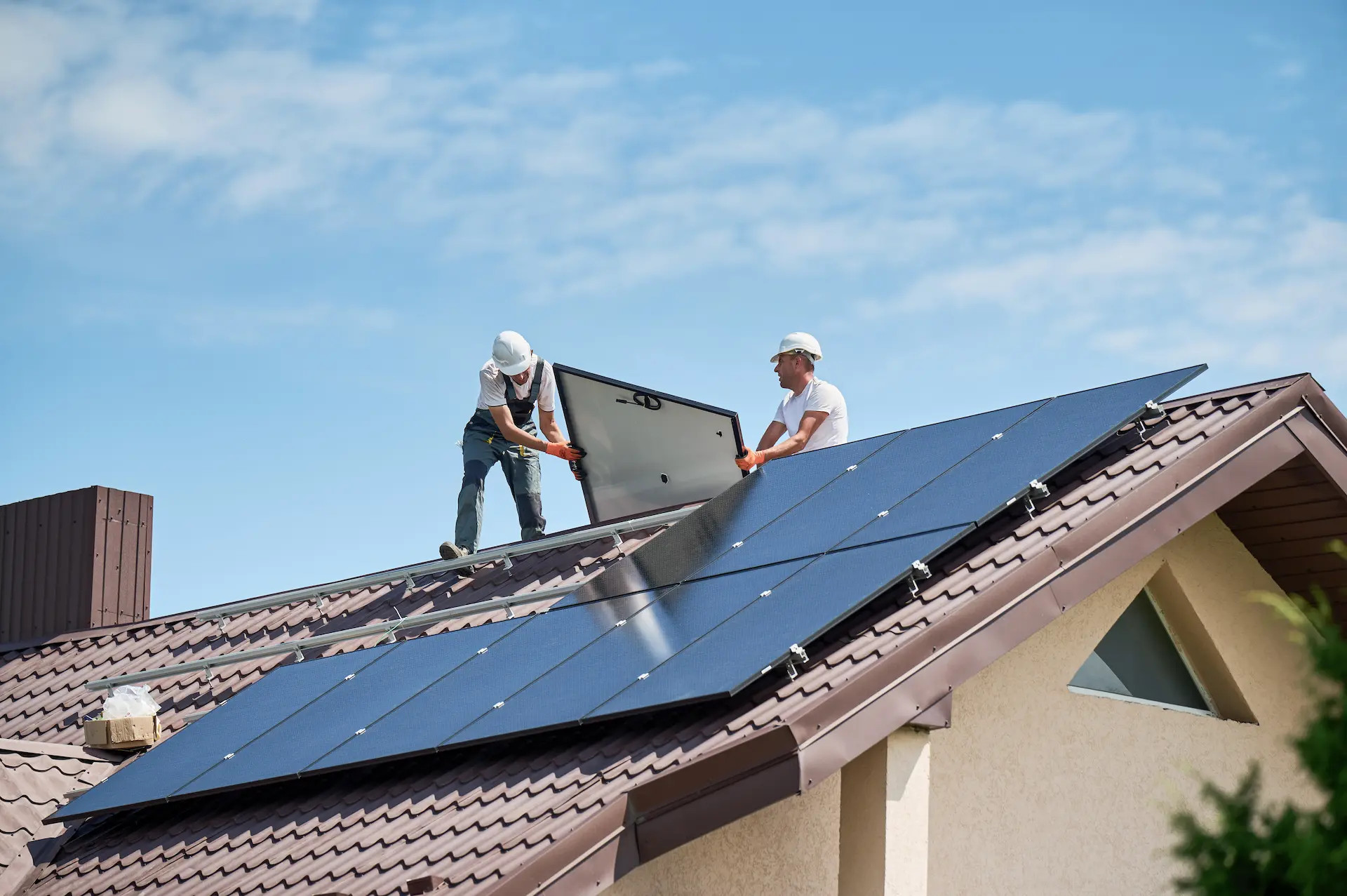 Zwei Arbeiter installieren Solarmodule auf einem Dach unter blauem Himmel.