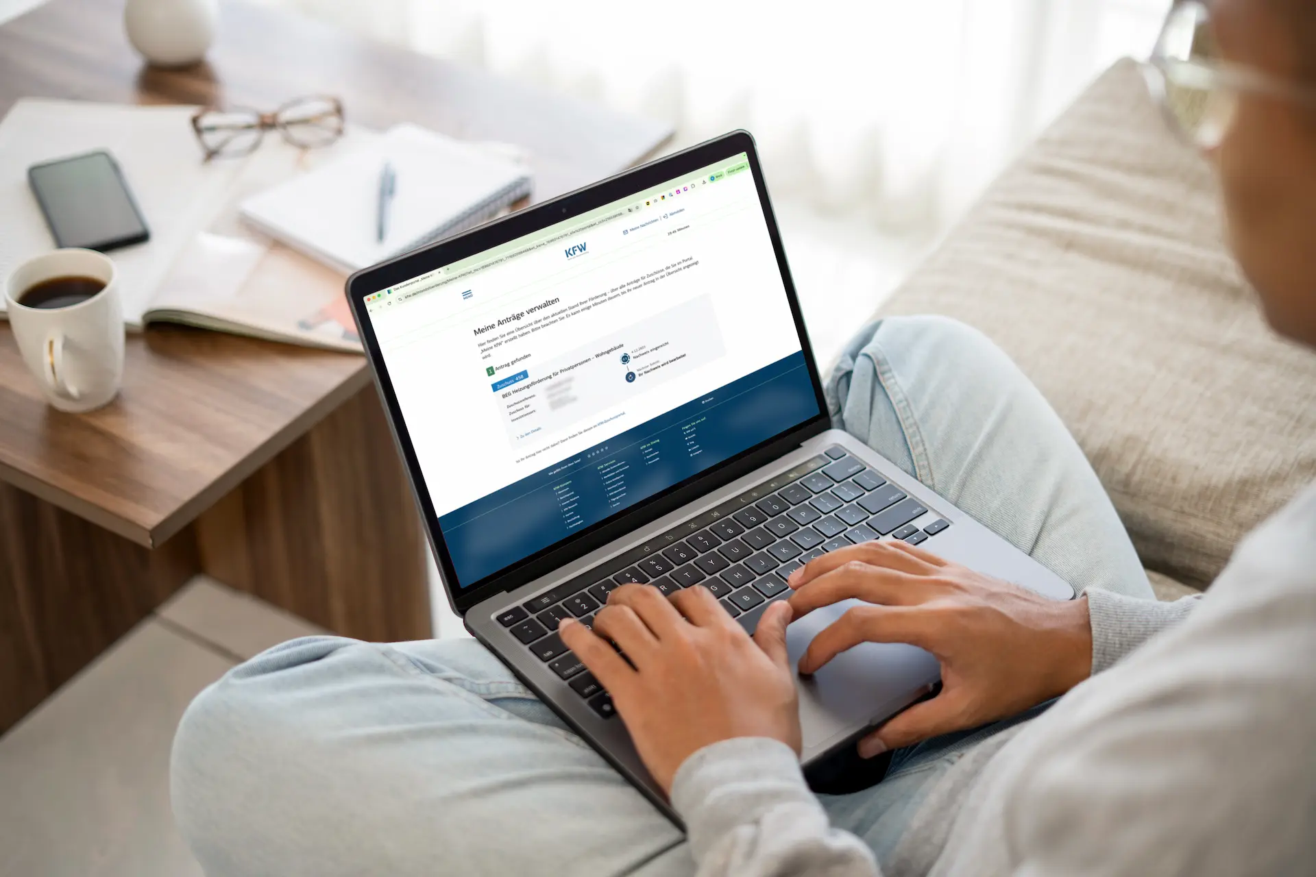 Person tippt auf einem Laptop mit einer KfW-Website zur Verwaltung von Förderanträgen, daneben Kaffeetasse, Smartphone und Notizbuch auf einem Holztisch.