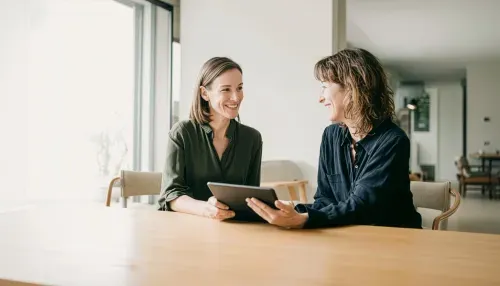 Zwei Frauen sitzen an einem Tisch und führen eine freundliche Beratung mit einem Tablet durch.