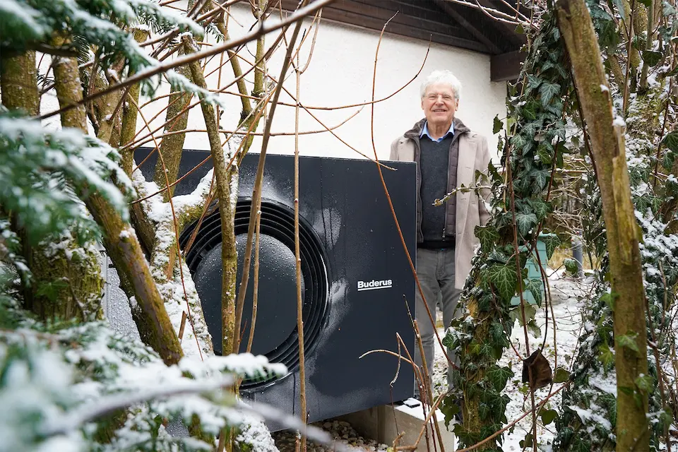 Älterer Mann steht im schneebedeckten Garten neben einer schwarzen Buderus Wärmepumpe vor einem Haus.