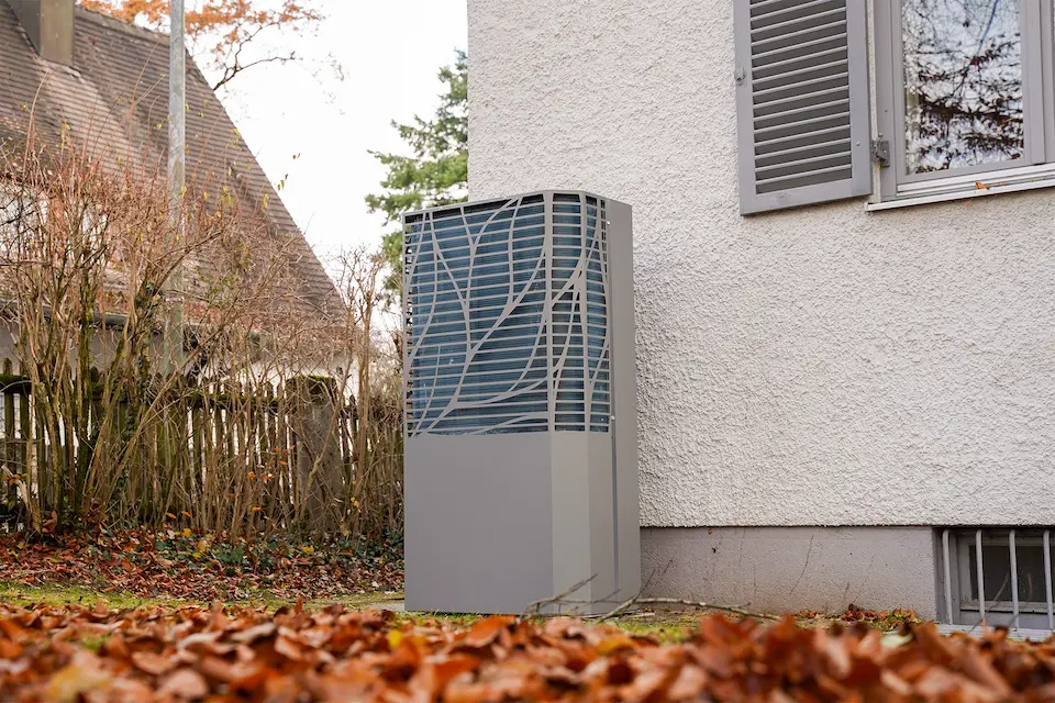 Moderne Wärmepumpe neben einem weißen Haus im Garten mit herbstlichen Blättern auf dem Boden.