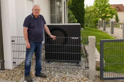 Älterer Mann steht neben einem schwarzen Außengerät einer Wärmepumpe vor einem Haus mit Garten.