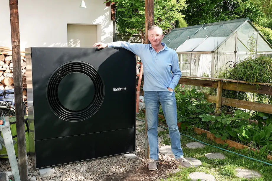 Älterer Mann lehnt an einer schwarzen Buderus-Heizungseinheit im Garten neben einem Gewächshaus.