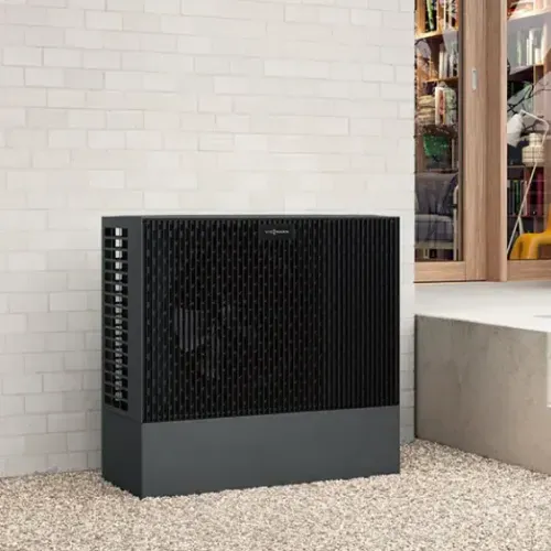 Schwarze Viessmann Wärmepumpe neben einem weißen Ziegelmauerwerk vor einem Gebäude mit großen Fenstern.