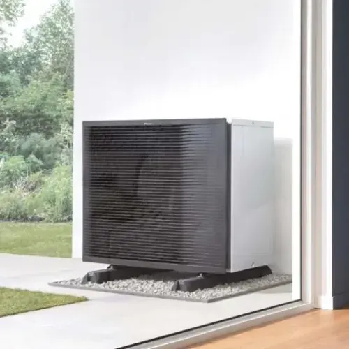 Außeneinheit einer modernen Klimaanlage oder Wärmepumpe auf Kies vor einem weißen Gebäudewand mit Garten im Hintergrund.