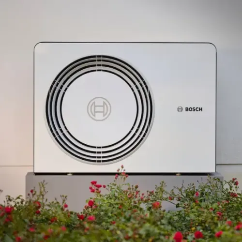 Weiße Bosch-Wärmepumpe auf einem grauen Sockel vor einer Wand, mit roten Blumen im Vordergrund.