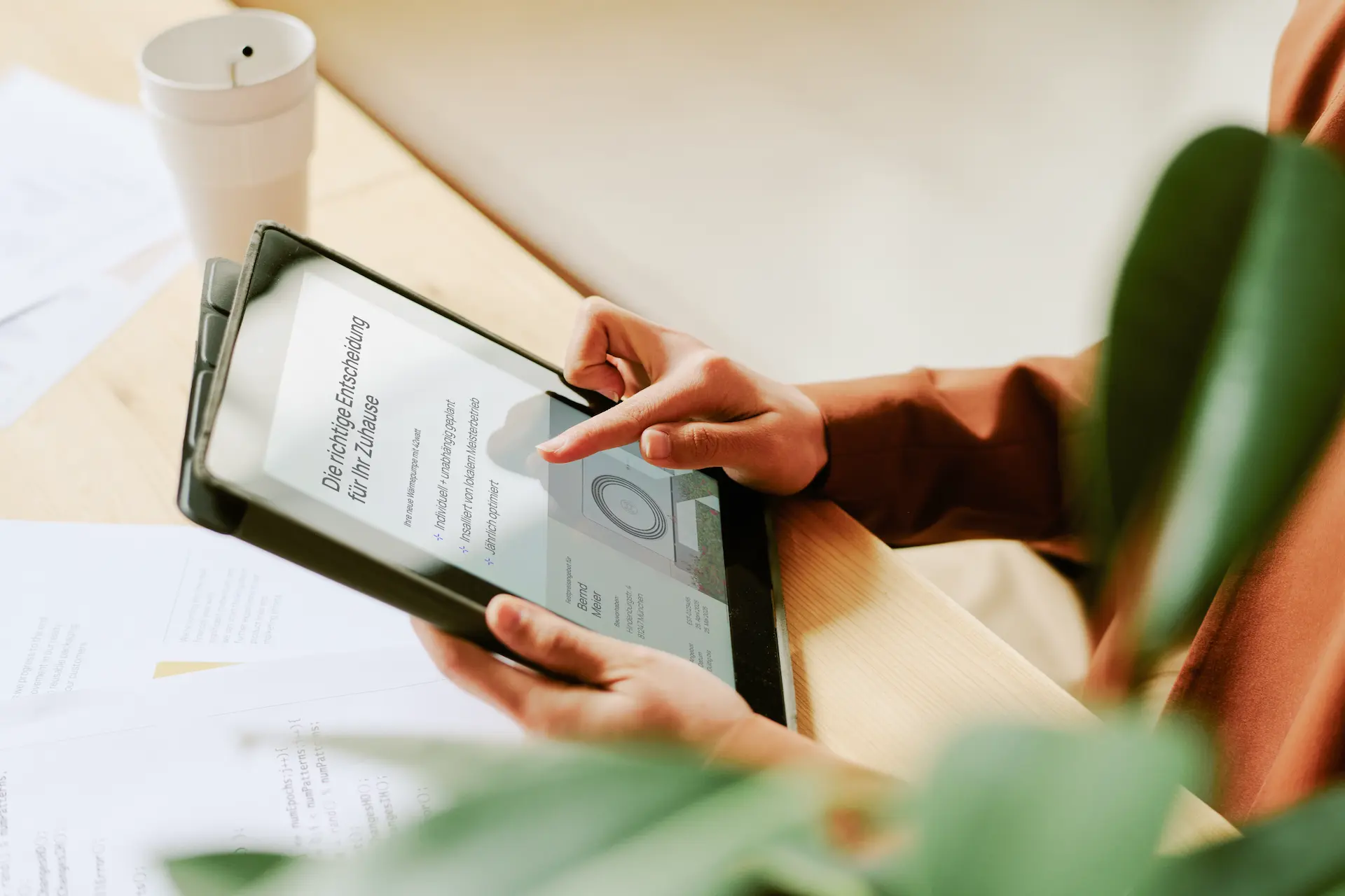 Person hält ein Tablet mit einer Anzeige über die richtige Entscheidung für das Zuhause, umgeben von Pflanzen und Papieren auf einem Holztisch.