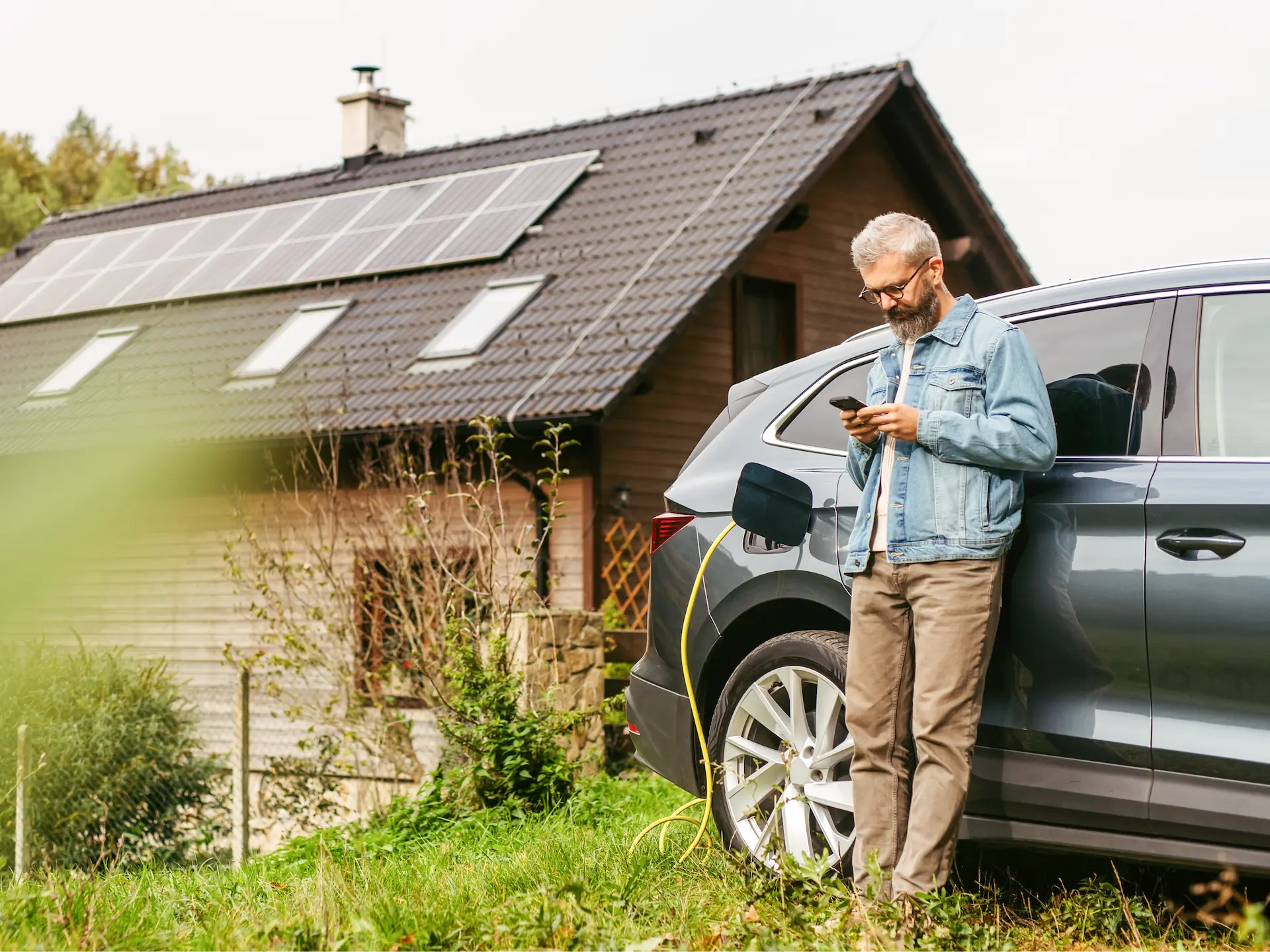 Mann steht neben einem Elektroauto, das an einem Haus mit Solarpanelen auf dem Dach aufgeladen wird, und schaut auf sein Smartphone.
