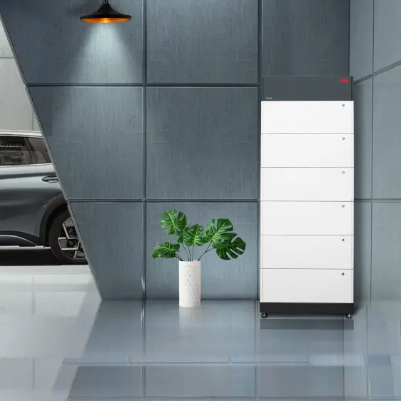 Modernes, weißes Energiespeichersystem in einem stilvollen Raum mit grauen Wänden und grünem Pflanzenarrangement neben einem grauen Auto.