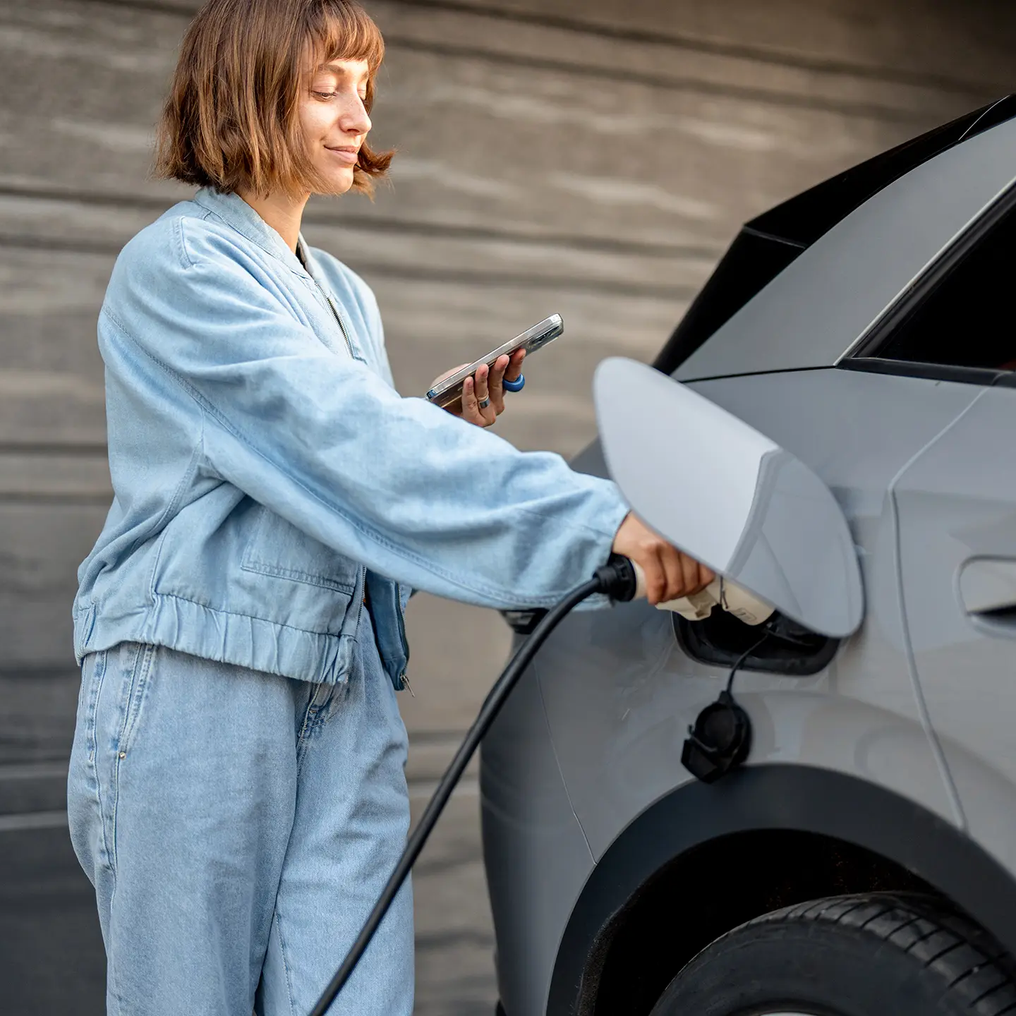 Frau lädt ihr graues Elektroauto und hält dabei ein Smartphone in der Hand.