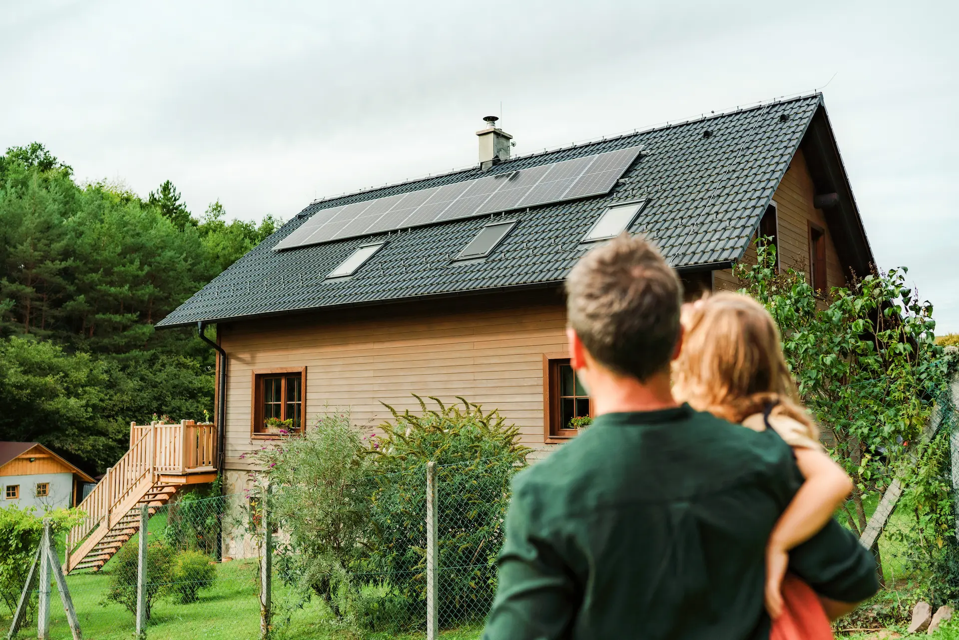 Ein Mann trägt ein Kind und schaut auf ein Haus mit Solarpaneelen auf dem Dach und einem Garten mit Bäumen und Zaun.