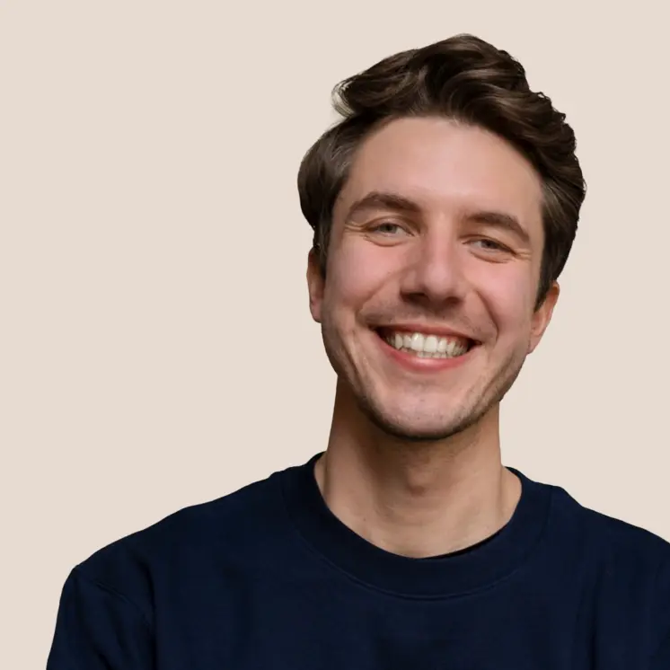 Junger Mann mit braunen Haaren lächelt vor einfarbigem beige Hintergrund.