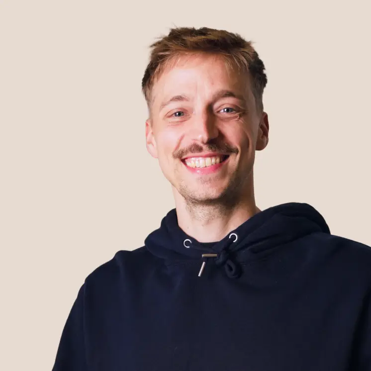 Junger Mann mit kurzem hellbraunem Haar, Schnurrbart und schwarzem Kapuzenpullover, der lächelt vor einem hellen Hintergrund.