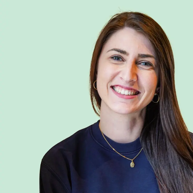 Lächende Frau mit langen braunen Haaren, Goldohrringen und Halskette vor hellem Hintergrund.