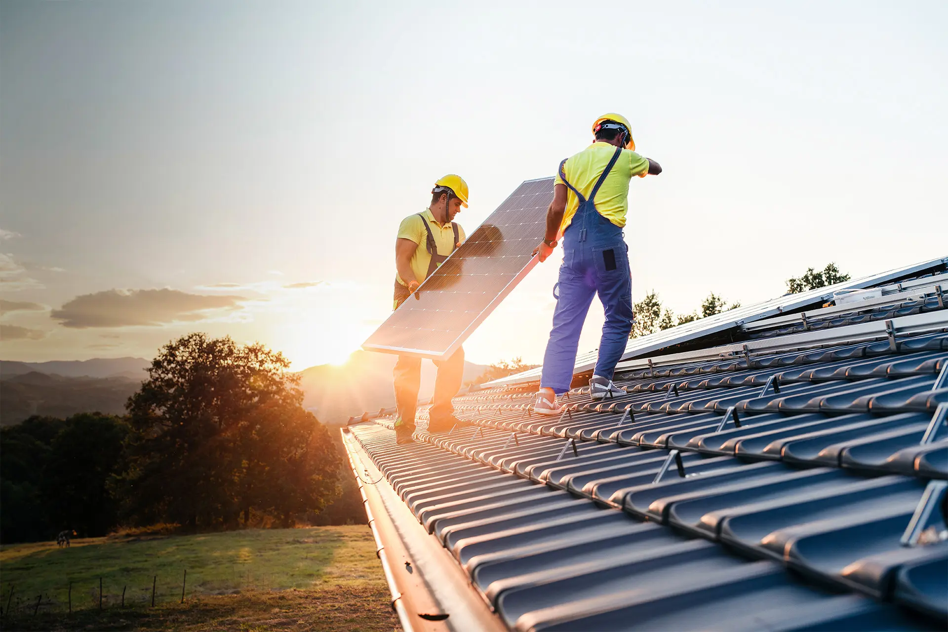 Zwei Arbeiter mit Schutzhelmen montieren eine Solaranlage auf einem Dach bei Sonnenuntergang.