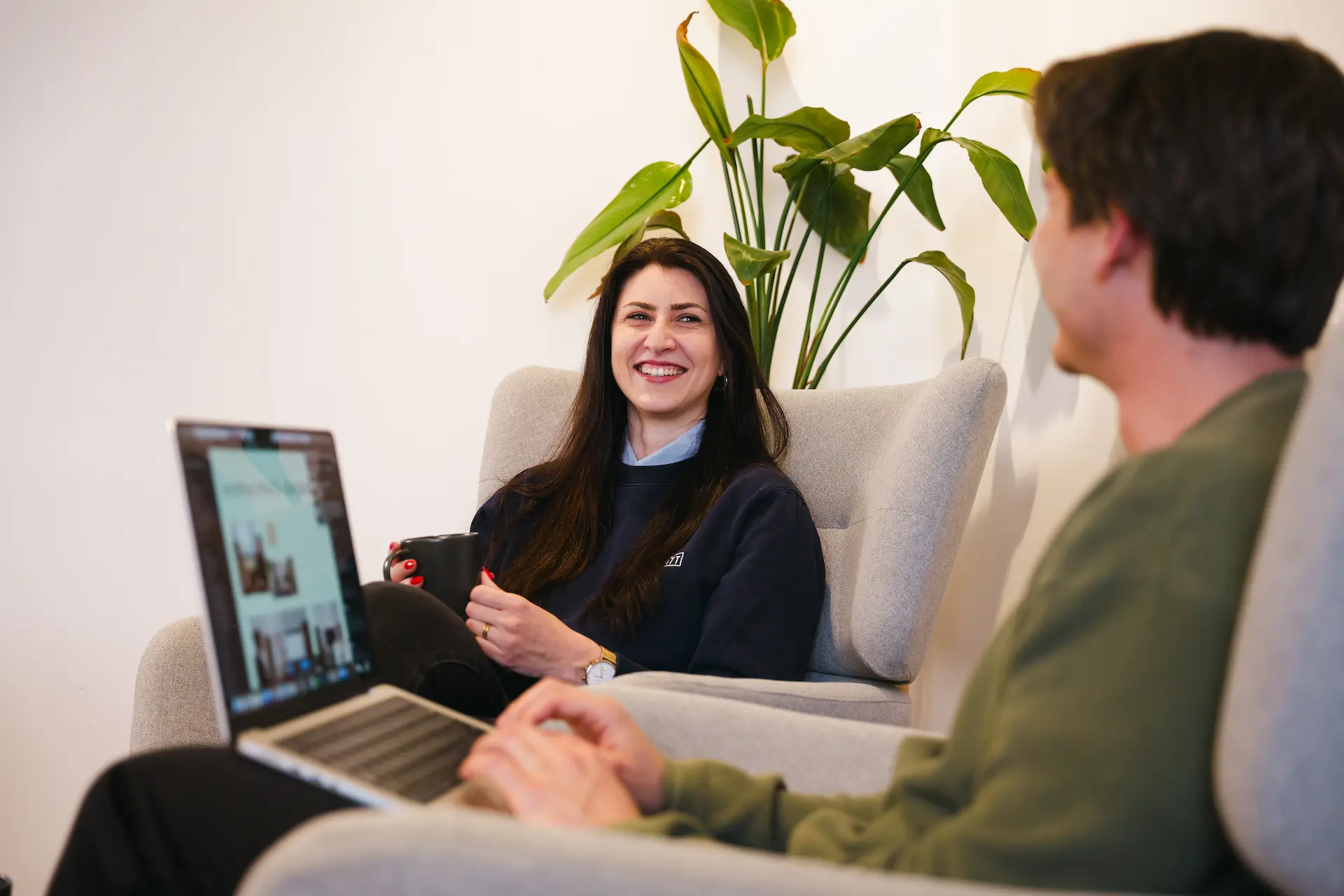 Zwei Personen unterhalten sich in einem gemütlichen Raum, eine Frau lächelt und hält eine Tasse, während der andere mit einem Laptop auf dem Schoß sitzt.