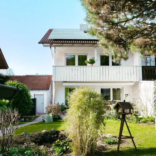 Weißes zweistöckiges Haus mit Balkon, Solarpaneelen auf dem Dach und Garten mit Vogelhaus und Pflanzen.