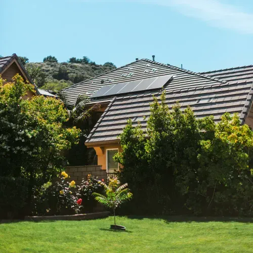 Hausdach mit Solarzellen und grüner Gartenlandschaft mit Büschen, Blumen und Rasen.