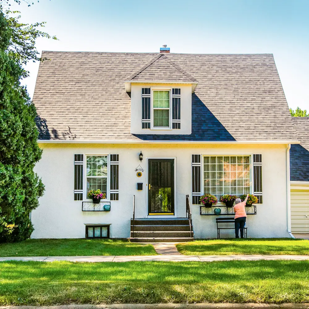 Ein Einfamilienhaus mit einem grauen Giebeldach und weißen Wänden; eine ältere Frau pflanzt Blumen in Blumenkästen unter dem großen Fenster.
