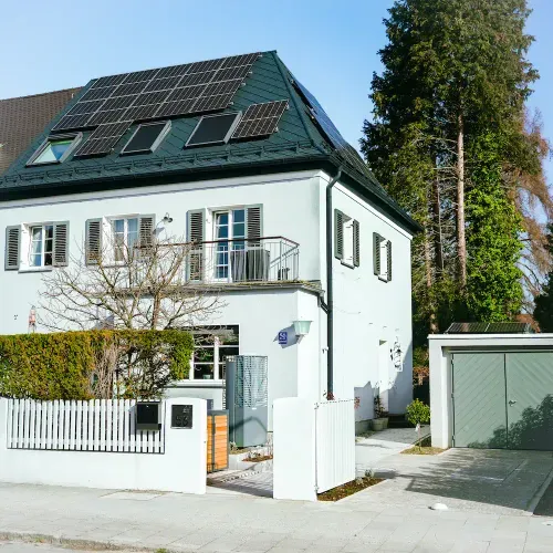 Zweistöckiges weißes Haus mit Solarpaneelen auf dem Dach, Balkon und angrenzender Garage, umgeben von Hecke und Bäumen.