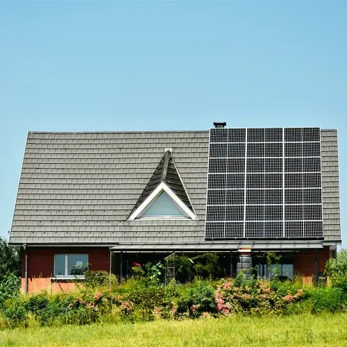 Haus mit grauem Schindeldach, auf dem rechteckige Solarmodule installiert sind, umgeben von grünem Gras und Blumen.