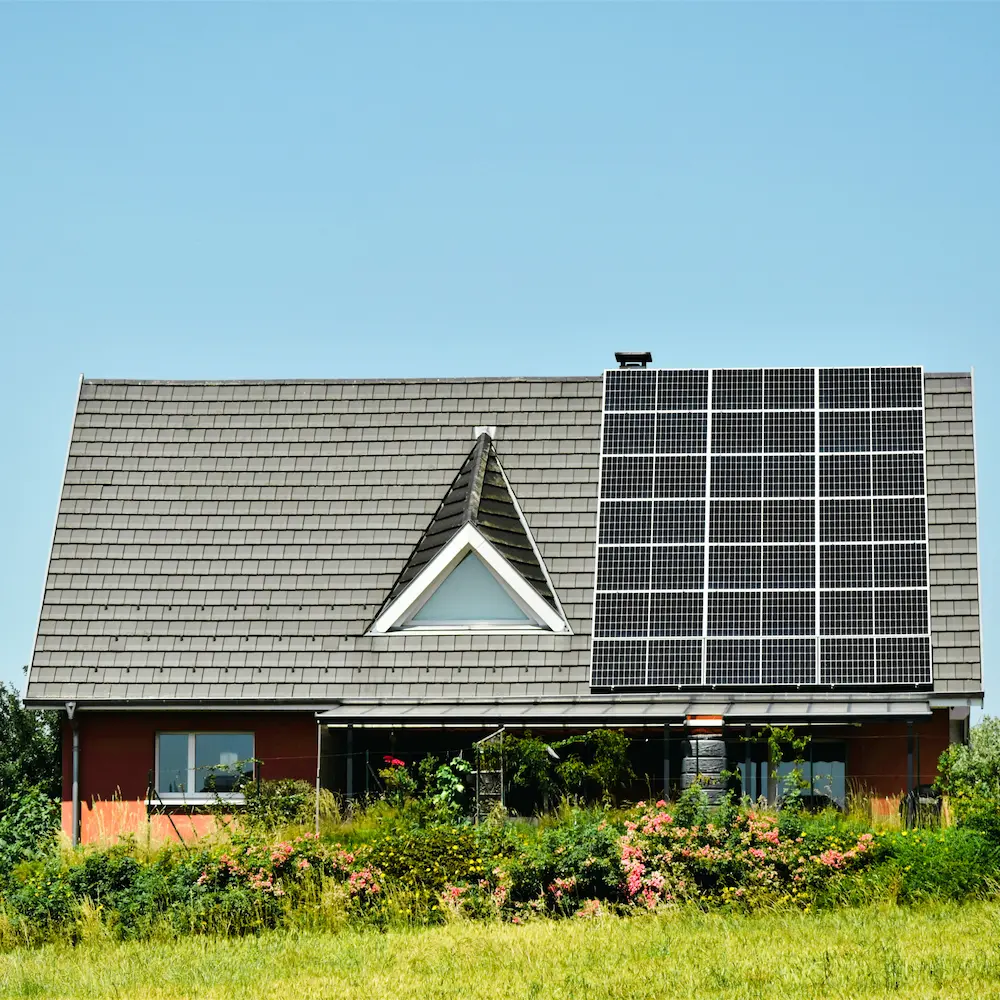 Haus mit grauem Schindeldach, auf dem rechteckige Solarmodule installiert sind, umgeben von grünem Gras und Blumen.