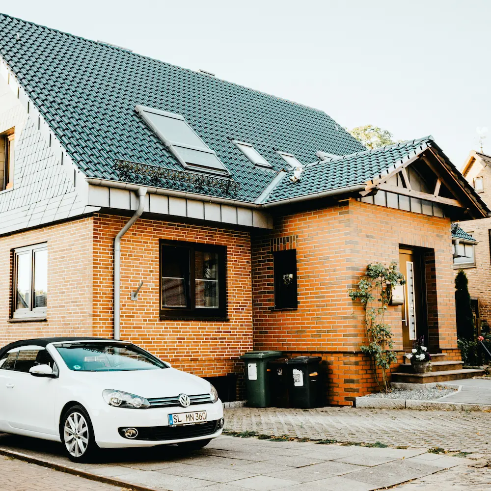 Ziegelsteinhaus mit grünem Ziegeldach und weißen Dachfenstern, davor ein weißes Volkswagen Cabrio auf der Straße.
