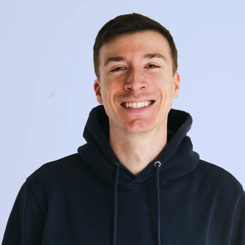 Junger Mann lächelt in schwarzer Kapuzenjacke vor hellblauem Hintergrund.