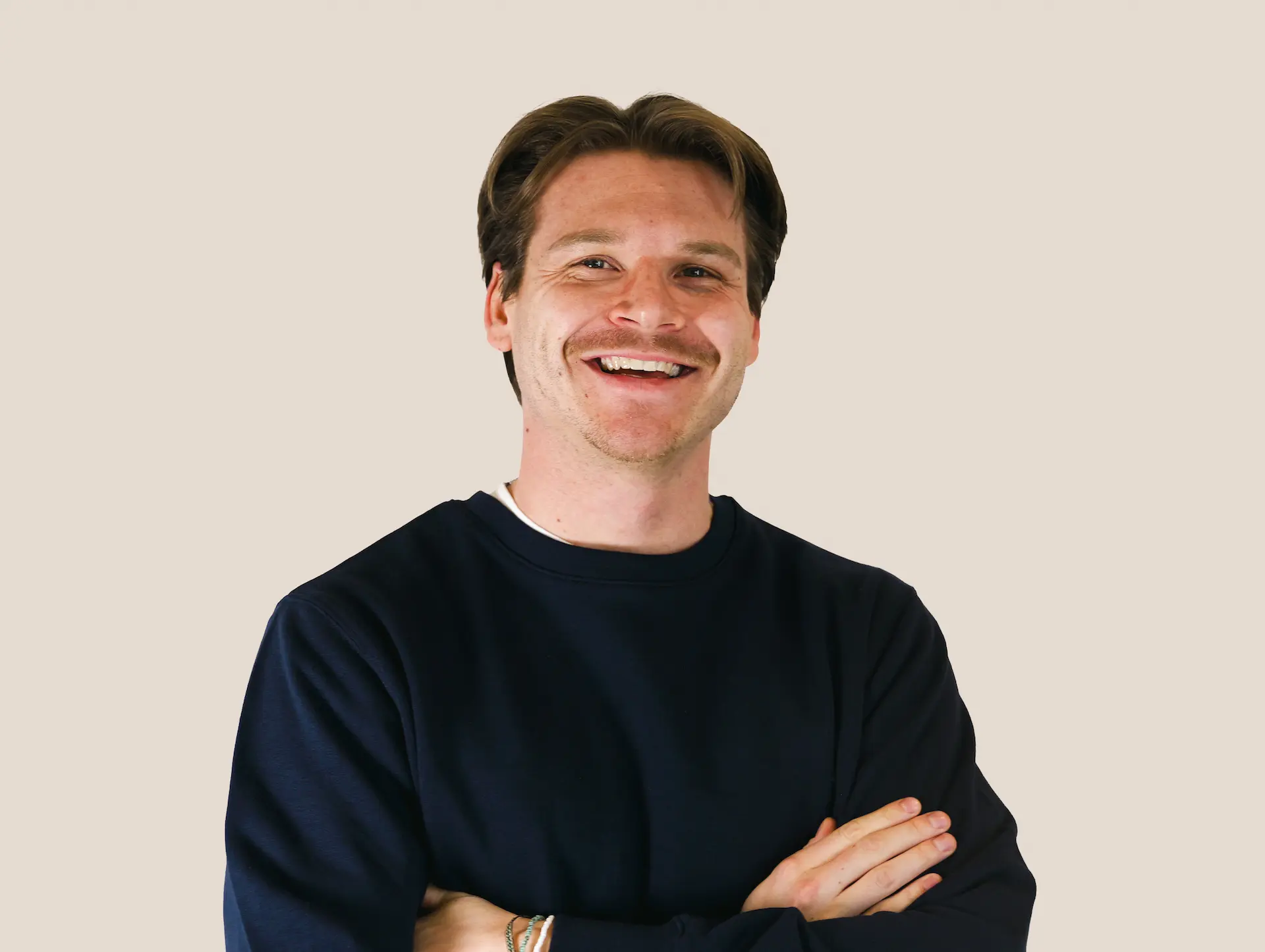 Lächelnder Mann mit braunen Haaren, dunklem Pullover und verschränkten Armen vor hellem Hintergrund.