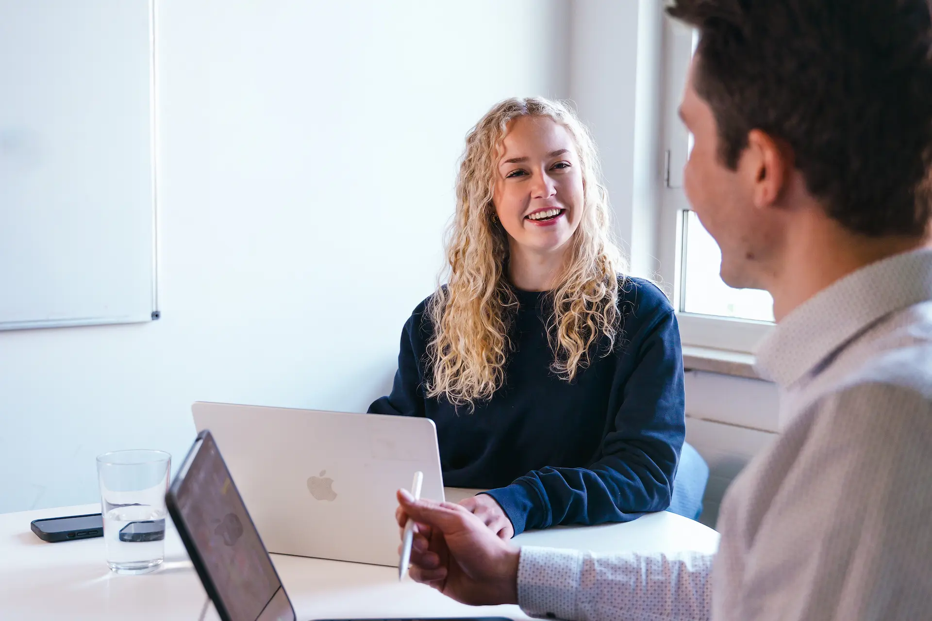 Zwei Personen unterhalten sich fröhlich an einem Tisch mit Laptops, eine Frau lächelt und ein Mann zeigt mit einem Stift.