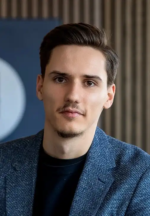 Porträt eines jungen Mannes mit kurzen dunklen Haaren und leichtem Bart, gekleidet in ein blaues Sakko über einem schwarzen Hemd vor unscharfem Hintergrund mit Streifen.