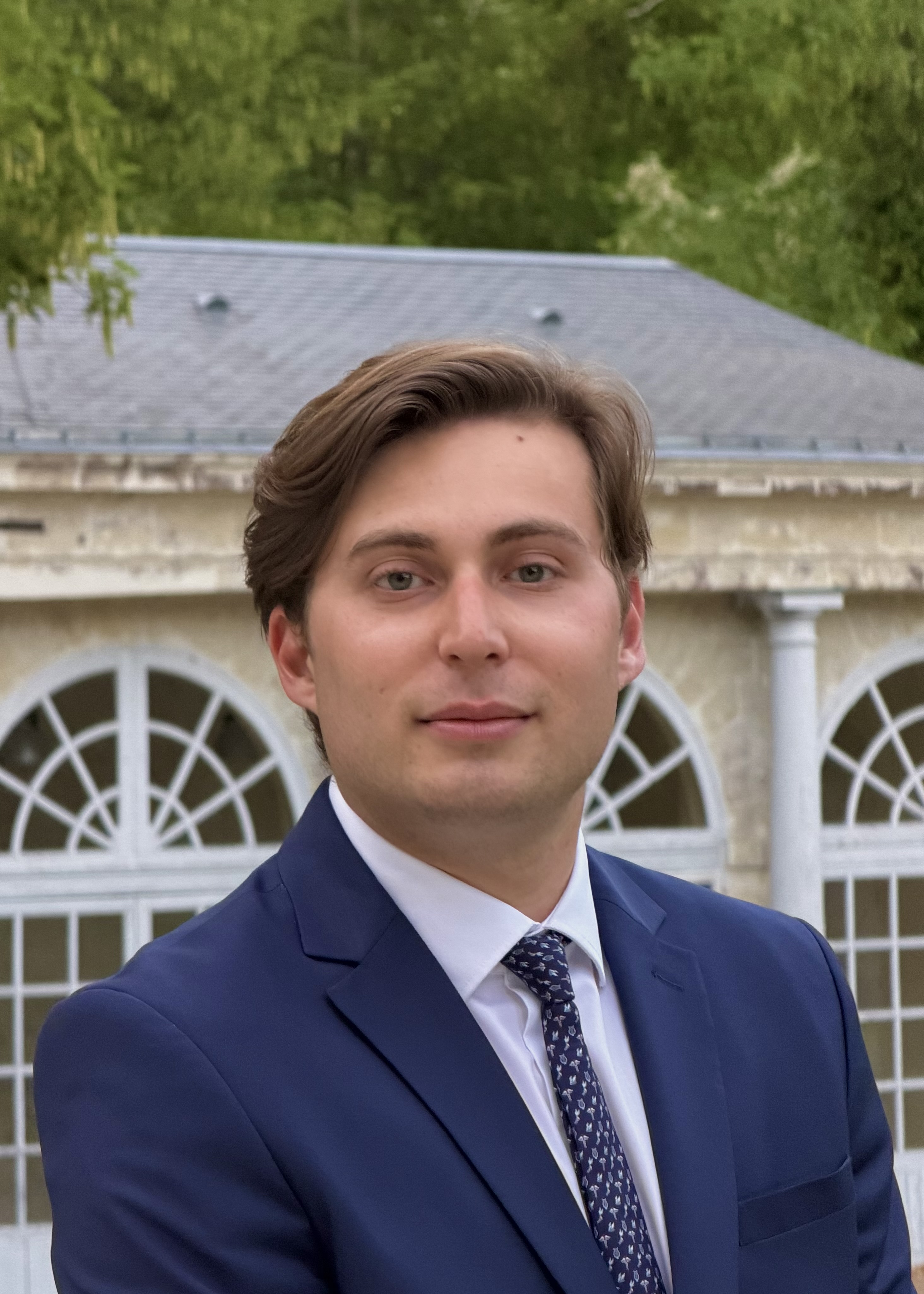 Young man with brown hair in a blue suit and patterned tie standing outdoors near building with arched windows.