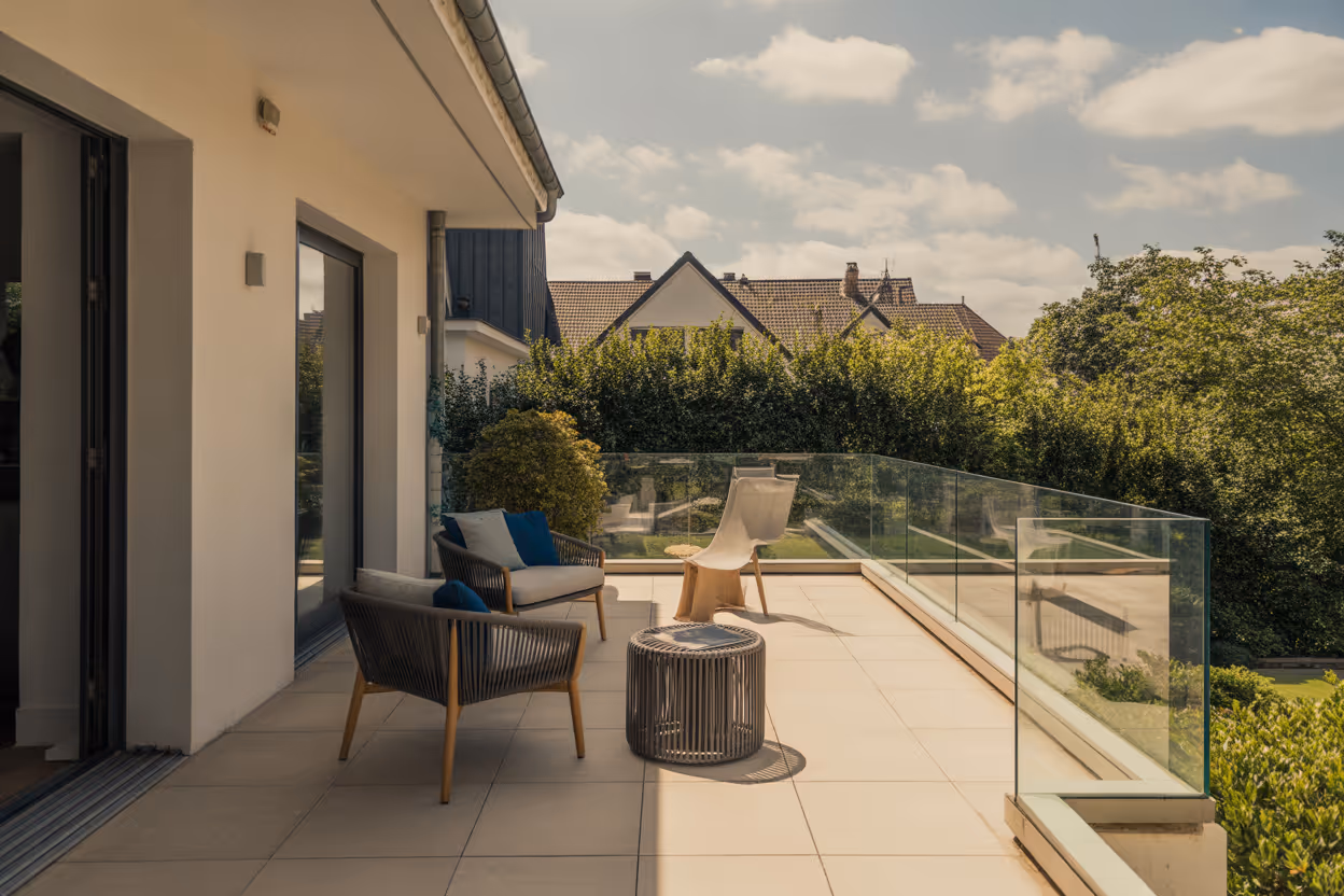 Moderne Plattenterrase mit zwei Sesseln, einem Stuhl, einem kleinen Tisch und Glasgeländer, umgeben von grünem Gebüsch und Hausdächern im Hintergrund.