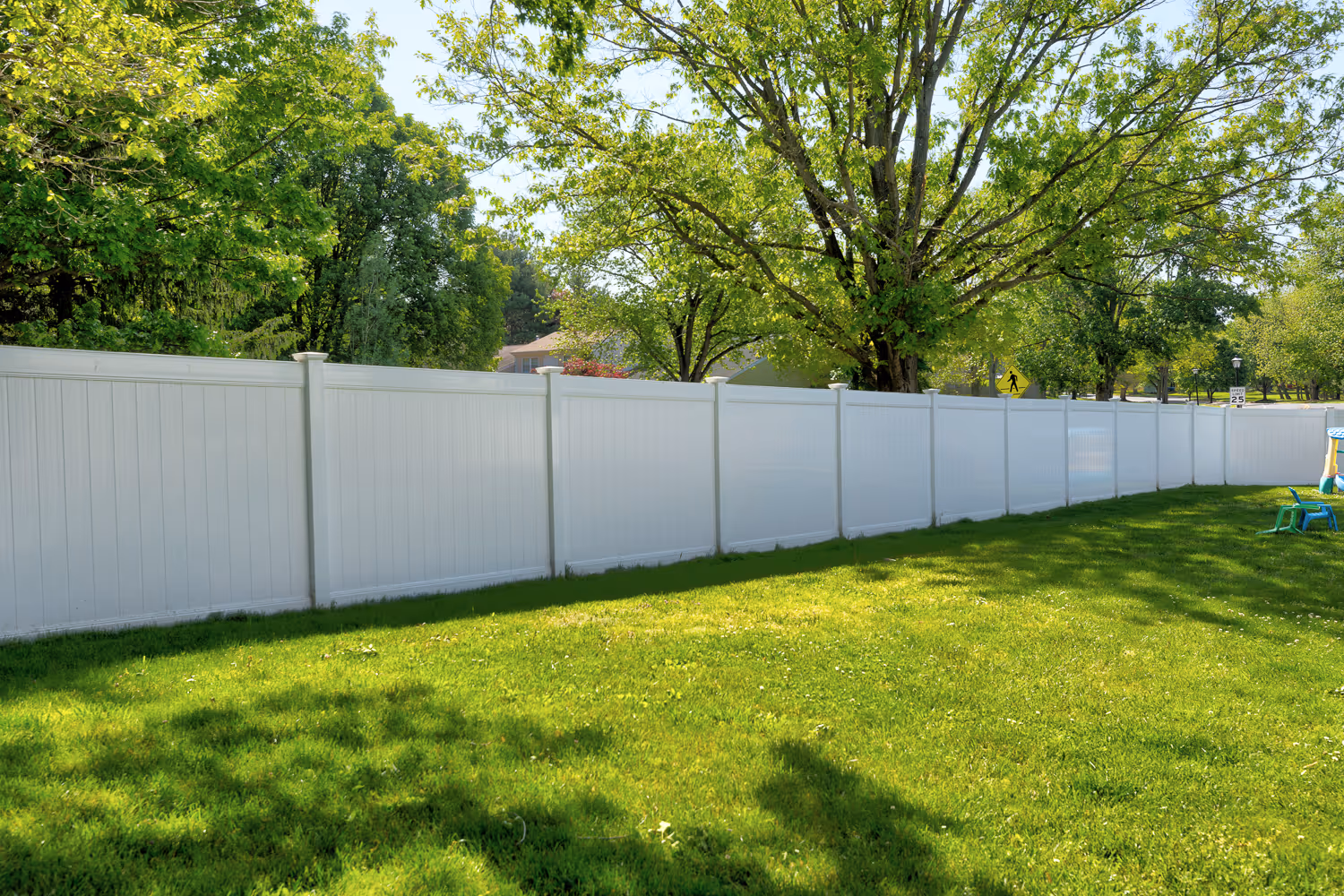 Long white vinyl fence in a sunny backyard with green grass and leafy trees behind it.