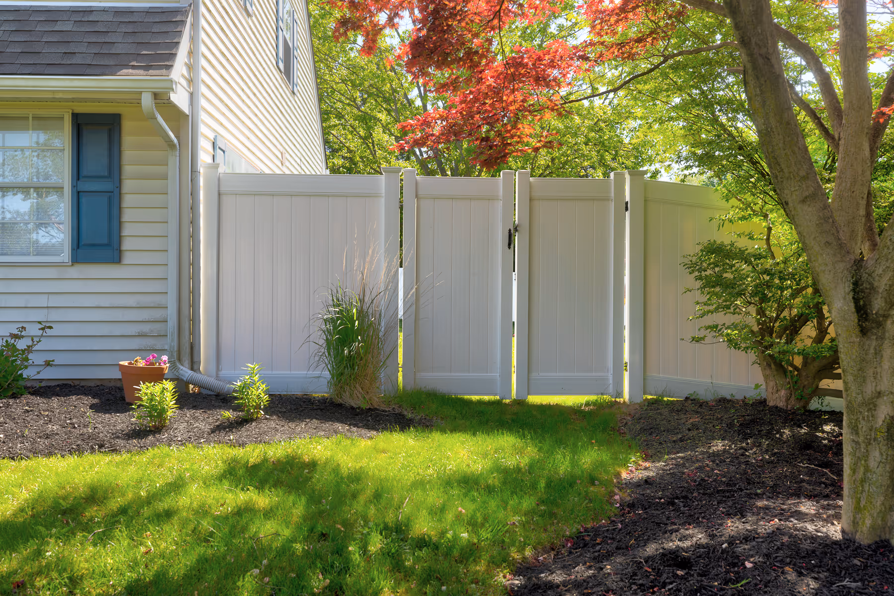 White vinyl privacy fence with gate beside the corner of a house and garden with grass and trees.