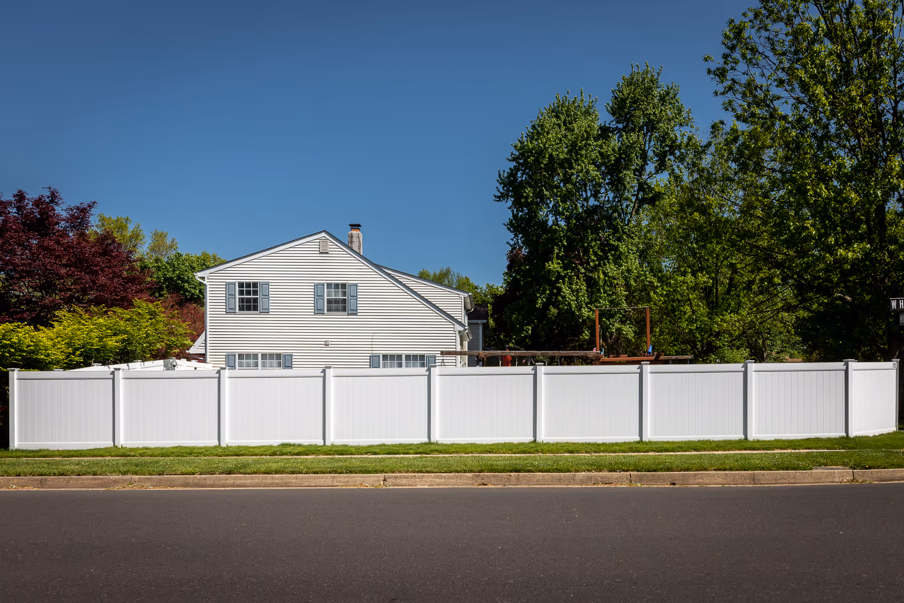 White vinyl privacy fence enclosing a backyard with a two-story house and green trees behind it.
