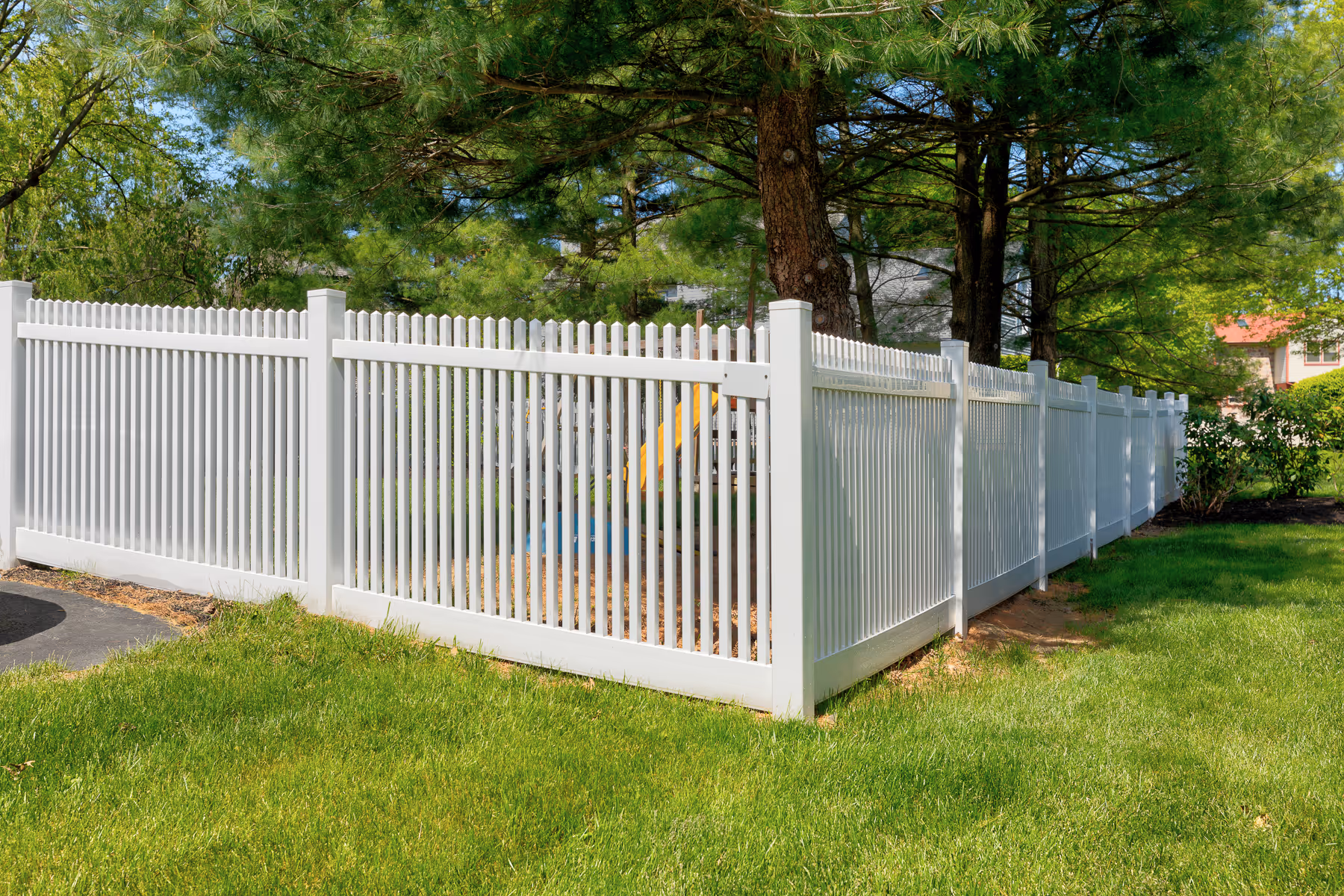 White vinyl picket fence enclosing a green yard with trees and playground equipment.