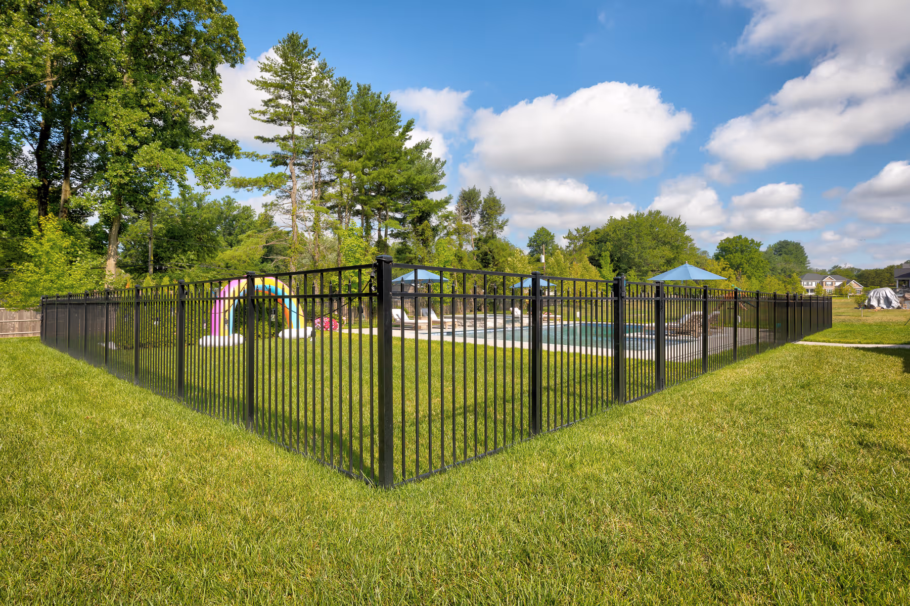 Black aluminum fence enclosing a backyard pool area with umbrellas and greenery under a blue sky.