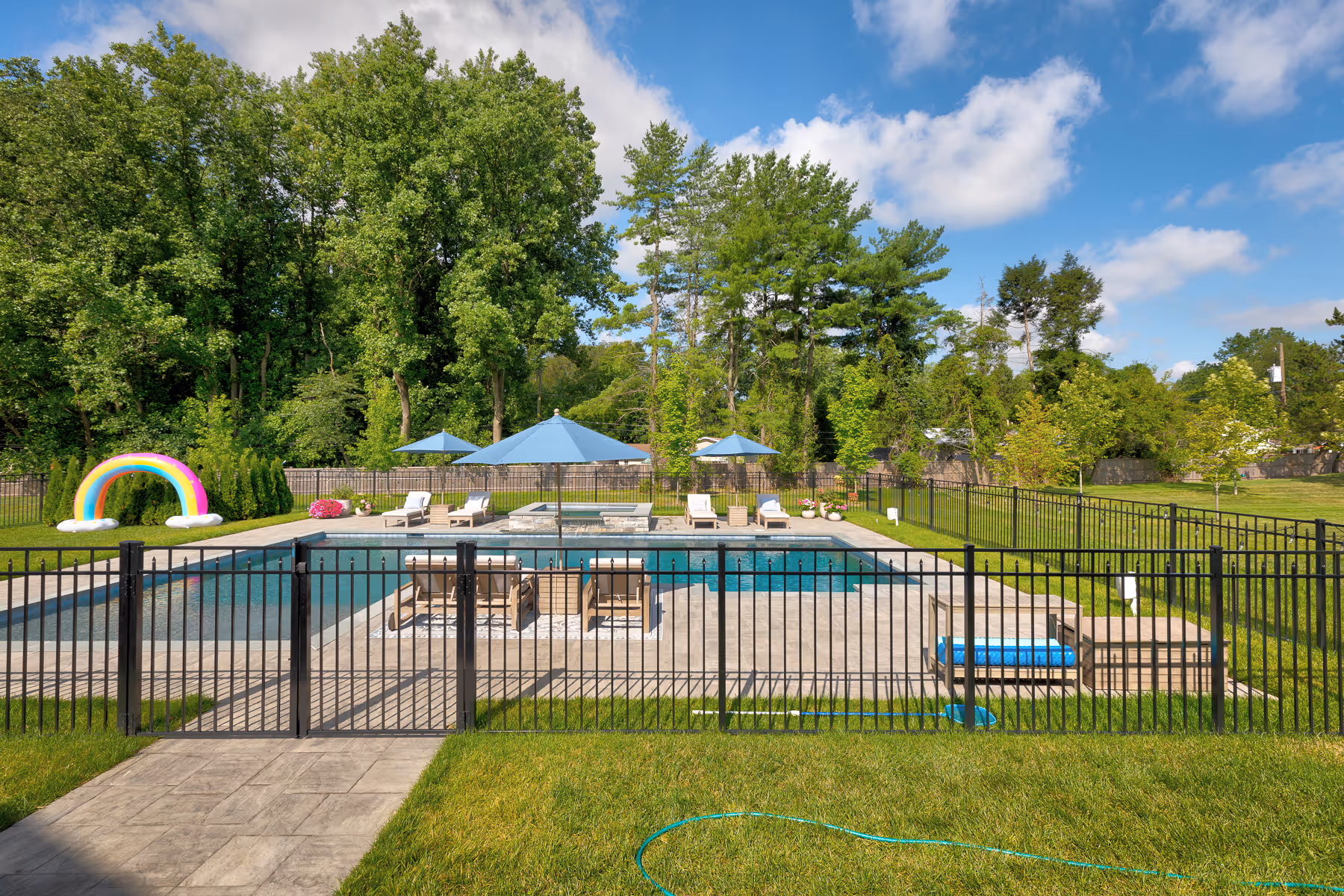 Black metal fence enclosing an outdoor pool area with chairs, umbrellas, and green trees beyond.