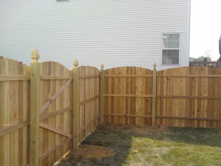 New wooden fence with arched tops enclosing a corner yard beside a white house.