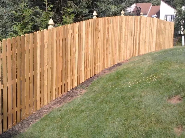 Long wooden fence with pointed tops running along a grassy slope near houses and trees.